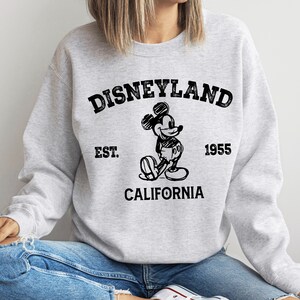 A woman wearing a gray sweatshirt with a graphic of Mickey Mouse and text that says "Disneyland Est. 1955 California".