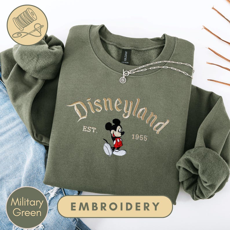 A military green sweatshirt with a Disney Mickey Mouse embroidered on it, along with a chain and a spool of thread, indicating that it is an embroidery piece.