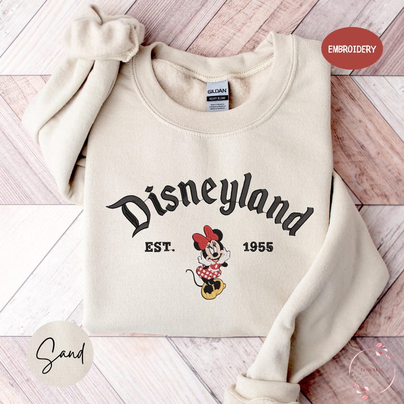 A beige sweatshirt with the text "Disneyland EST. 1955" and a Minnie Mouse graphic printed on it, laid out on a wooden surface.