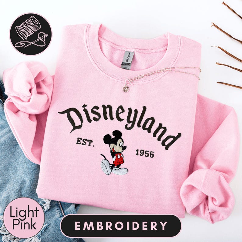 A pink sweatshirt with the text "Disneyland EST. 1955" and an embroidered image of Mickey Mouse, a well-known Disney character.
