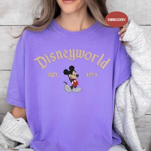 A woman wearing a purple t-shirt with a Mickey Mouse design and text that says "Disneyworld EST. 1971".