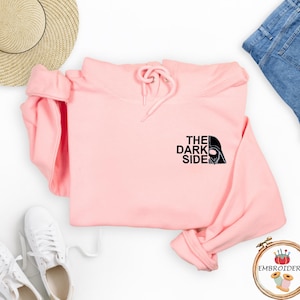 May include: A light pink hoodie with the text "THE DARK SIDE" and a Darth Vader graphic. The hoodie is folded, with a drawstring hood and long sleeves. White sneakers, a straw hat, and blue jeans are also visible.