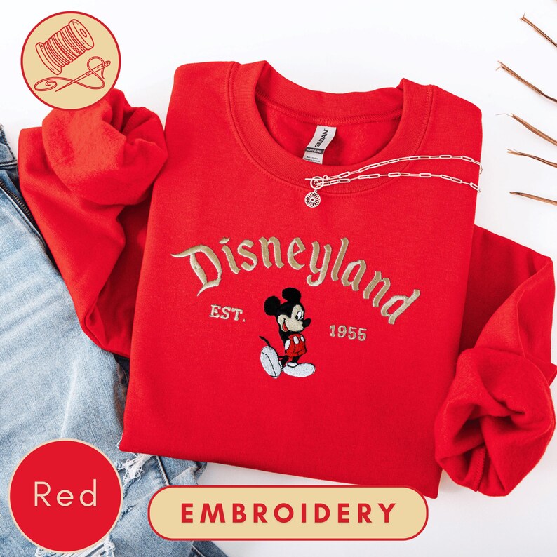 A red sweater with the Disneyland logo and the year 1955 embroidered on it, along with a pair of jeans and some thread.