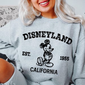A woman wearing a gray sweatshirt with a graphic of Mickey Mouse and text that says "DISNEYLAND EST. 1955 CALIFORNIA".