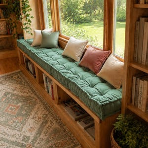 May include: A long, tufted sage green bench cushion sits atop a wooden window seat with built-in bookshelves. The cushion is accented with five square throw pillows in shades of cream, sage green, and dusty rose, arranged along the window frame.