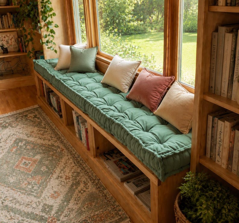 May include: A long, tufted sage green bench cushion sits atop a wooden window seat with built-in bookshelves. The cushion is accented with five square throw pillows in shades of cream, sage green, and dusty rose, arranged along the window frame.