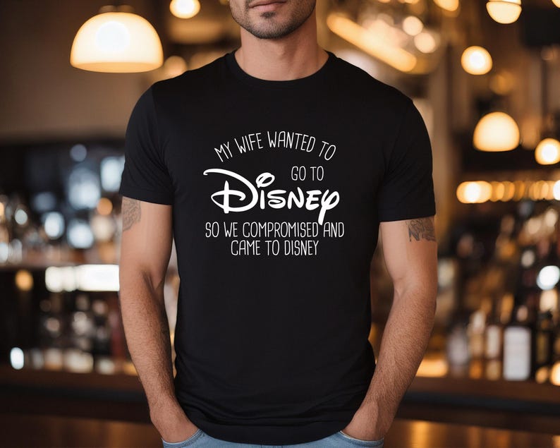 A man wearing a black t-shirt with white text that says "My wife wanted to go to Disney so we compromised and came to Disney".