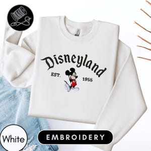 A white sweatshirt with the text "Disneyland EST. 1955" and an embroidered image of Mickey Mouse, a classic Disney character.