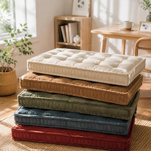 May include: A stack of five rectangular floor cushions in cream, tan, olive green, dark blue, and deep red. Each cushion features a tufted top surface with a grid pattern and vertical side panels, creating a structured, thick, and plush appearance.