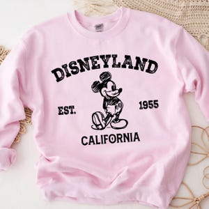 A pink sweatshirt with a graphic of Mickey Mouse and text that says "Disneyland Est. 1955 California".