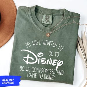 a green t-shirt with white text, laid out on a white surface. The text on the t-shirt reads "My wife wanted to go to Disney so we compromised and came to Disney". There is also a brown hat and a necklace visible in the image.