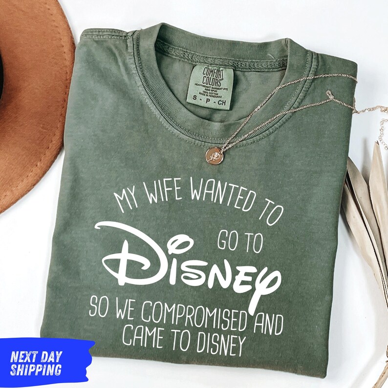 a green t-shirt with white text, laid out on a white surface. The text on the t-shirt reads "My wife wanted to go to Disney so we compromised and came to Disney". There is also a brown hat and a necklace visible in the image.