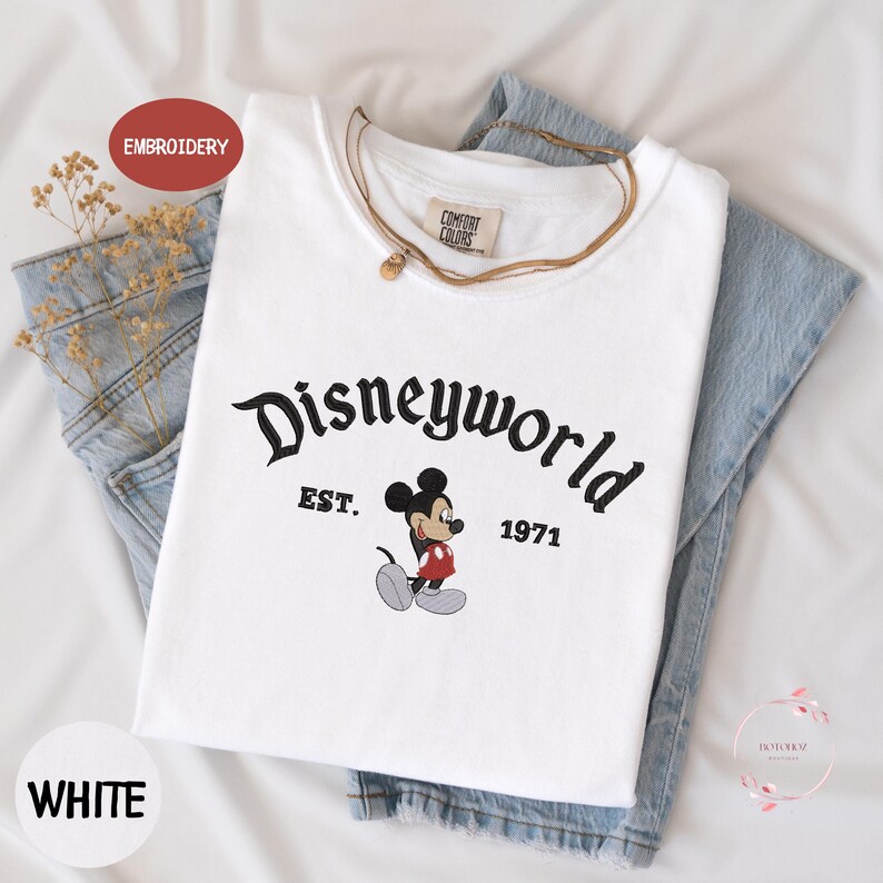 A white t-shirt with the text "Disneyworld EST. 1971" printed on it, along with a Mickey Mouse graphic. The t-shirt is placed on top of a pair of denim jeans.