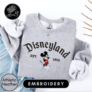 A gray sweatshirt with the text "Disneyland EST. 1955" and a Mickey Mouse embroidered on it, along with a spool of thread and a needle, suggesting the image is an embroidery of a Disney-themed sweatshirt.