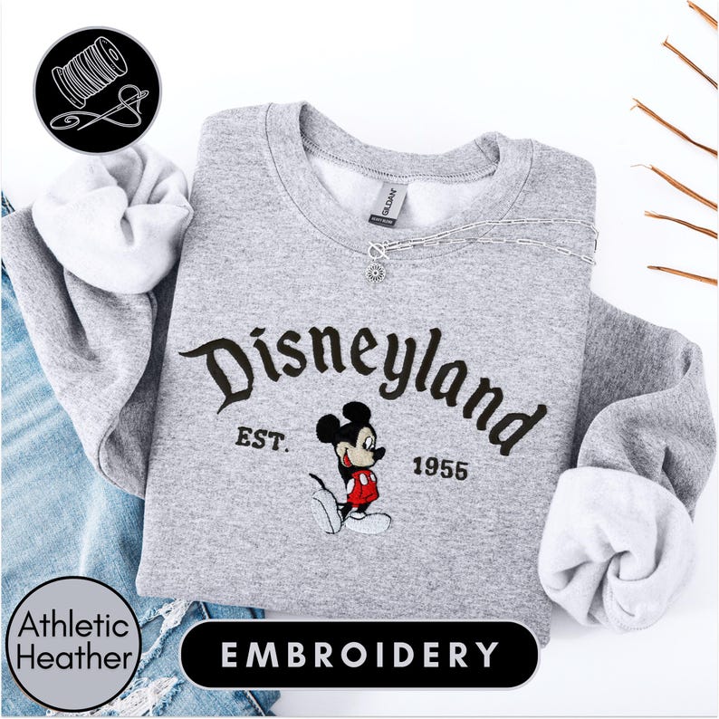 A gray sweatshirt with the text "Disneyland EST. 1955" and a Mickey Mouse embroidered on it, along with a spool of thread and a needle, suggesting the image is an embroidery of a Disney-themed sweatshirt.