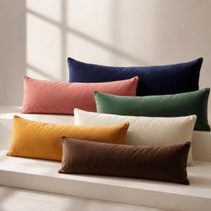 May include: A collection of six rectangular lumbar cushions in various solid colours including navy blue, dusty rose, forest green, cream, mustard yellow, and dark brown. Each cushion features a smooth, soft-touch fabric finish and a long, slim shape.