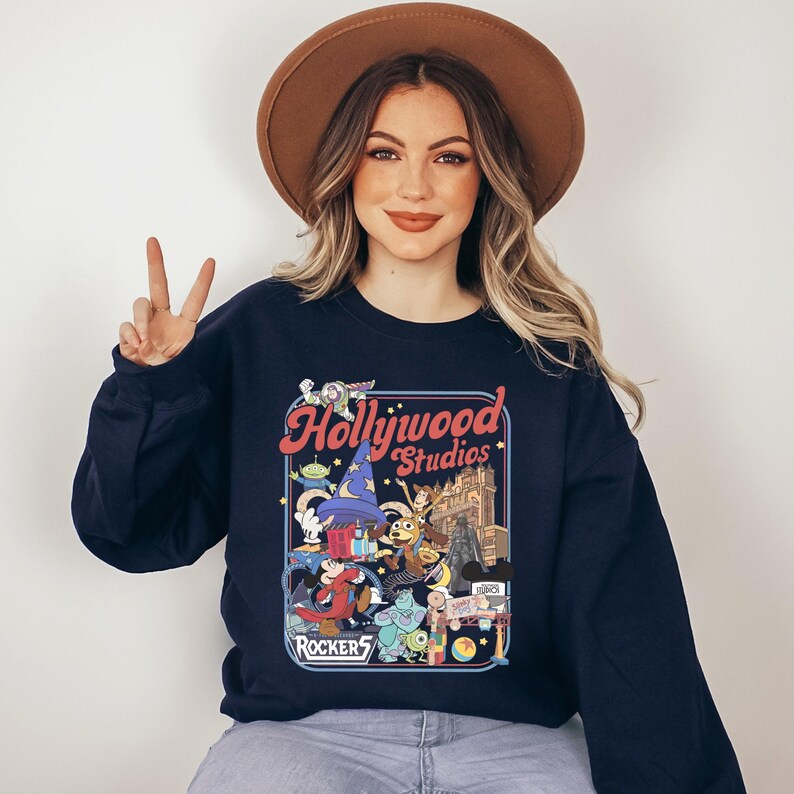 A woman wearing a black sweatshirt with a graphic of Disney characters and the text "Hollywood Studios" is making a peace sign.