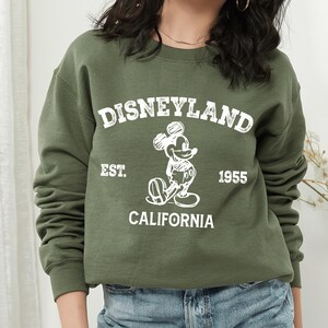 A woman wearing a green sweatshirt with the text "Disneyland Est. 1955" and a Mickey Mouse graphic printed on it.