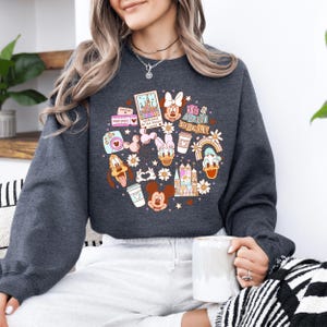 May include: Dark grey sweatshirt with a circular design featuring Disney characters and the text "It's a small world!". The design includes Mickey Mouse, Minnie Mouse, Donald Duck, Daisy Duck, Goofy, and a rainbow. The sweatshirt is a comfortable, casual style.