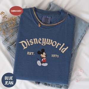 a blue Disneyworld t-shirt with the text "Disneyworld EST. 1971" and a Mickey Mouse graphic, along with a denim jacket and jeans.