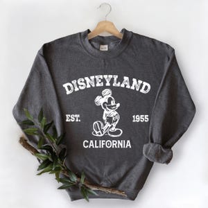 A gray sweatshirt with a graphic of Mickey Mouse and text that says "Disneyland Est. 1955 California" hanging on a wooden hanger.