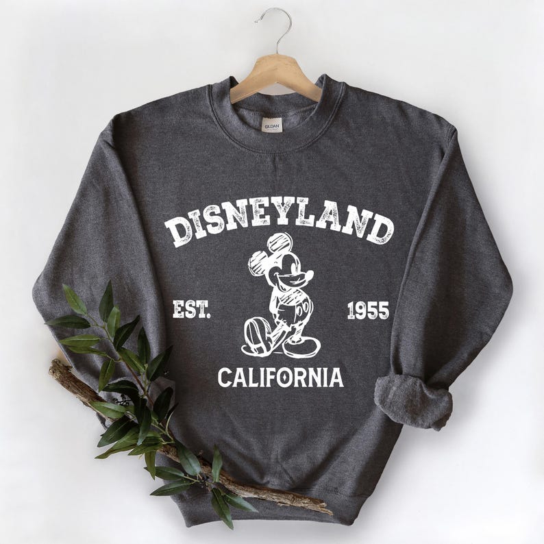 A gray sweatshirt with a graphic of Mickey Mouse and text that says "Disneyland Est. 1955 California" hanging on a wooden hanger.