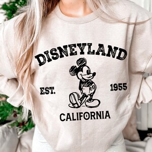 A person wearing a white sweatshirt with a graphic of Mickey Mouse and text that says "Disneyland Est. 1955 California".