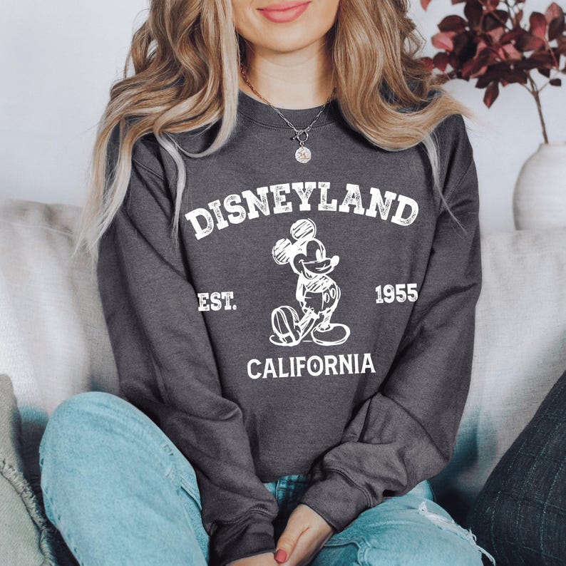 A woman wearing a gray sweatshirt with the text "Disneyland EST. 1955 CALIFORNIA" printed on it, sitting on a couch.