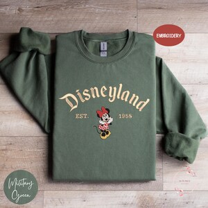 A green sweatshirt with the text "Disneyland EST. 1955" and an embroidered Minnie Mouse character, laid out on a wooden surface.