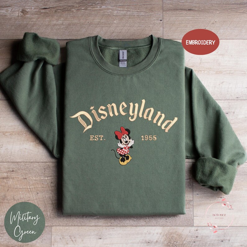 A green sweatshirt with the text "Disneyland EST. 1955" and an embroidered Minnie Mouse character, laid out on a wooden surface.