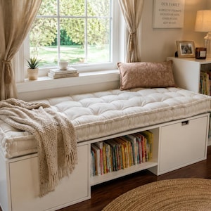 May include: A white rectangular storage bench featuring a tufted off-white cushion on top, a beige knit throw blanket draped over one side, and open shelving filled with books. The bench includes two closed storage compartments with handles.