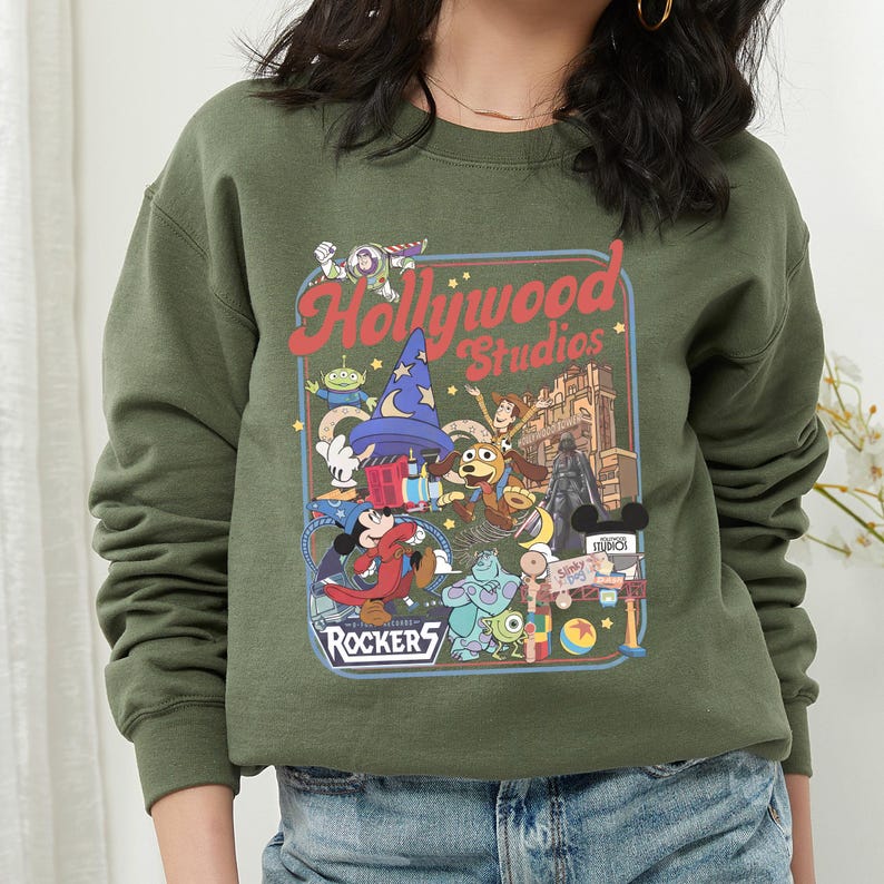 A woman wearing a green sweatshirt with a colorful graphic design featuring various Disney characters and logos, including Mickey Mouse, Minnie Mouse, and the Rocker's logo.