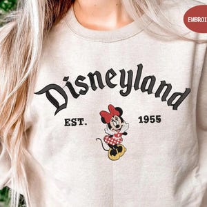 A person wearing a beige sweatshirt with the text "Disneyland EST. 1955" and a Minnie Mouse graphic printed on it.