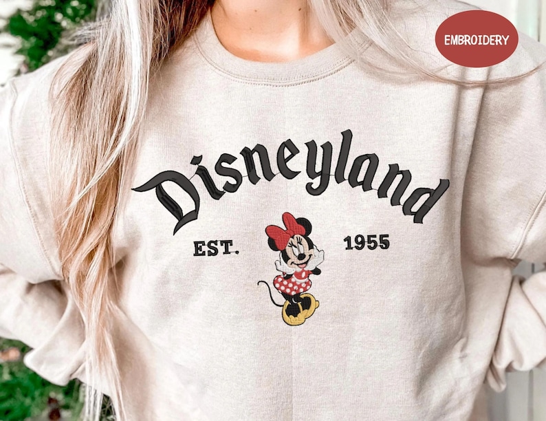 A person wearing a beige sweatshirt with the text "Disneyland EST. 1955" and a Minnie Mouse graphic printed on it.