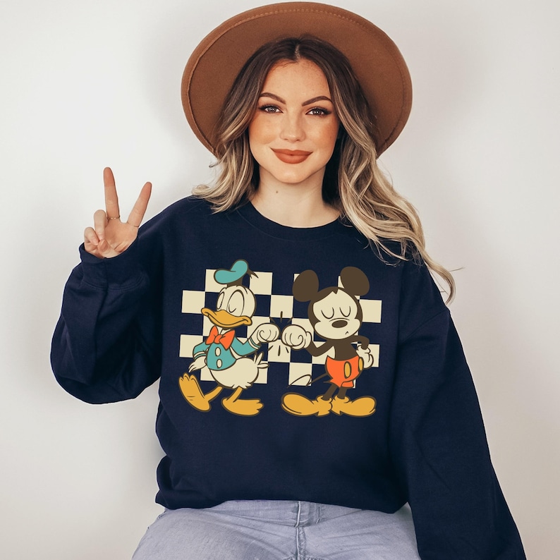 A woman wearing a black sweatshirt with a cartoon character design and a brown hat is making a peace sign.