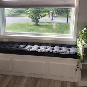 May include: A long, rectangular dark gray tufted bench cushion positioned on a white built-in window seat. The cushion features a series of evenly spaced button tufts across its surface, providing a textured appearance beneath a large window.
