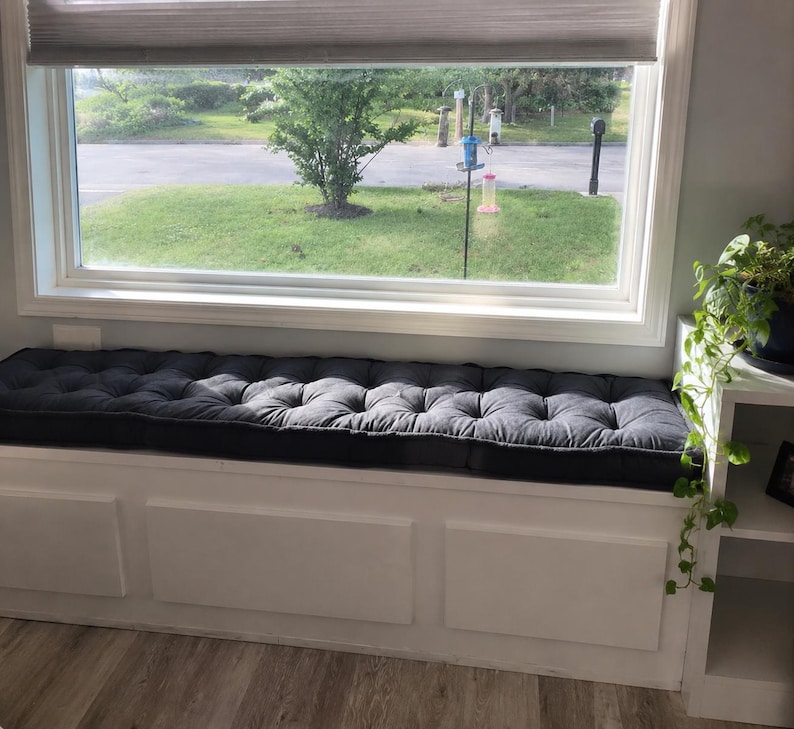 May include: A long, rectangular dark gray tufted bench cushion positioned on a white built-in window seat. The cushion features a series of evenly spaced button tufts across its surface, providing a textured appearance beneath a large window.