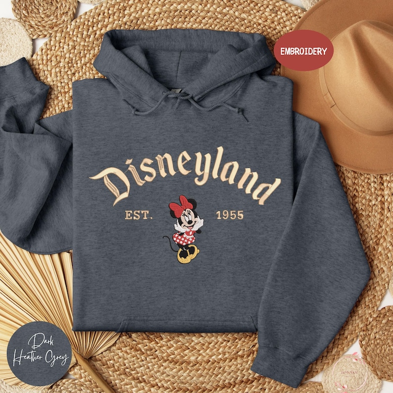 A gray hoodie with the text "Disneyland EST. 1955" and a Minnie Mouse embroidered on it, along with a straw hat and a woven mat in the background.