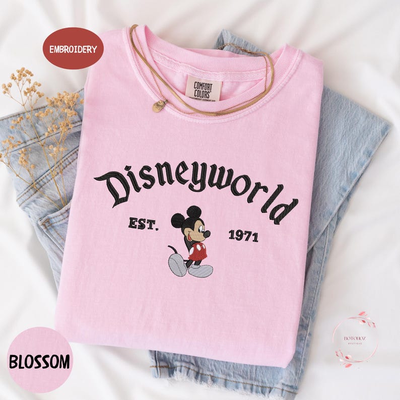 A pink t-shirt with the text "Disneyworld EST. 1971" printed on it, along with a Mickey Mouse graphic. The t-shirt is placed on top of a pair of blue jeans.