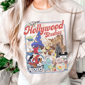 A person wearing a white sweatshirt with a colorful graphic design featuring various Disney characters and landmarks, including Mickey Mouse, Minnie Mouse, and the Eiffel Tower.