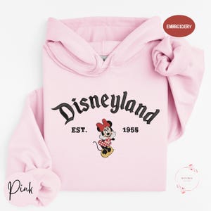 A pink hoodie with the text "Disneyland EST. 1955" and a Minnie Mouse graphic, indicating it is a souvenir or merchandise item from the Disney theme park.