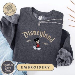 a gray sweatshirt with a Disney Mickey Mouse embroidered on it, along with a denim jacket and a pair of jeans. The sweatshirt has the text "Disneyland EST. 1955" and "Heather Dark Gray" printed on it.