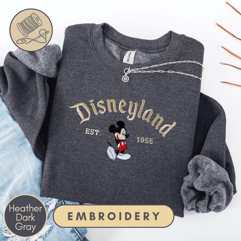 a gray sweatshirt with a Disney Mickey Mouse embroidered on it, along with a denim jacket and a pair of jeans. The sweatshirt has the text "Disneyland EST. 1955" and "Heather Dark Gray" printed on it.