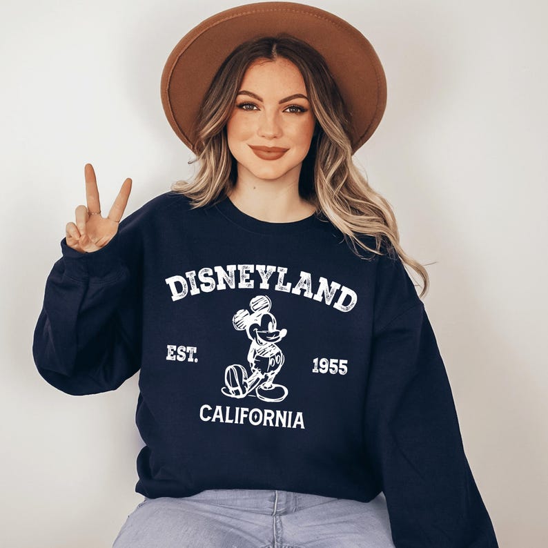 A woman wearing a black sweatshirt with a Disneyland logo and text, smiling and making a peace sign gesture.