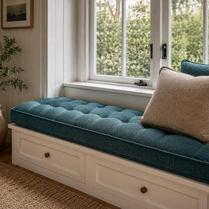May include: A white built-in window bench with two drawers featuring small round knobs. The bench has a tufted teal cushion and a single beige square throw pillow. The base is constructed with clean lines and a smooth white finish.