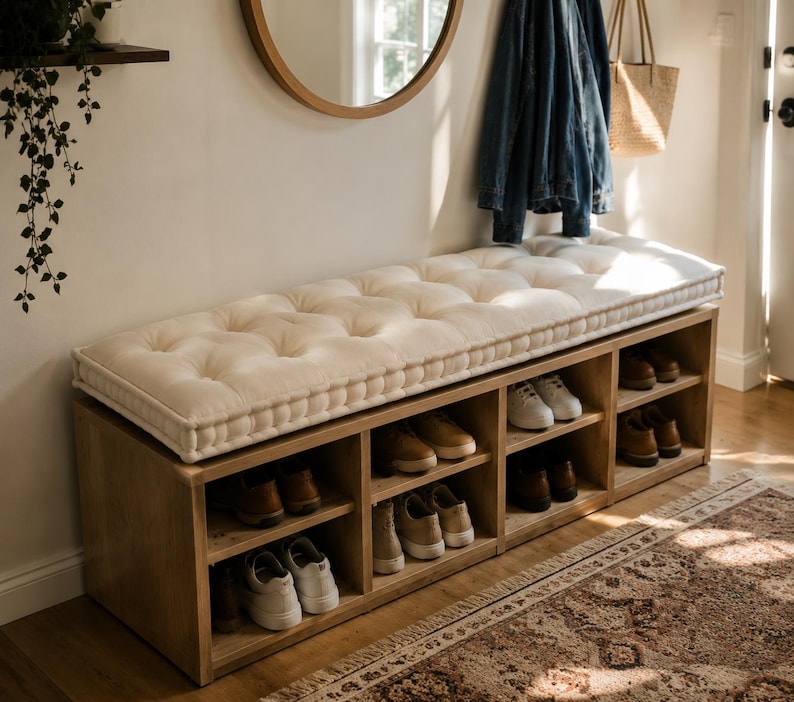 May include: A rectangular wooden shoe storage bench featuring a tufted off-white cushion on top. The base consists of eight open cubby compartments, each containing various pairs of sneakers and dress shoes in shades of white, tan, and brown.