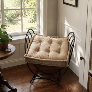 May include: A black metal stool with a curved frame and a thick, tufted beige cushion. The seat features a square shape with button detailing, supported by a rounded, open-work metal base that curves upward to form side handles.