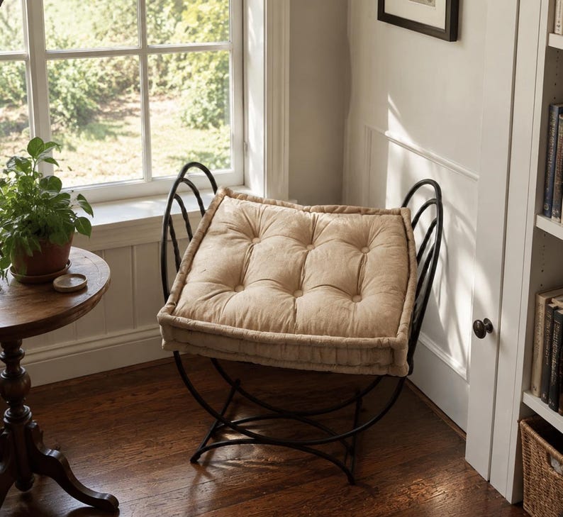 May include: A black metal stool with a curved frame and a thick, tufted beige cushion. The seat features a square shape with button detailing, supported by a rounded, open-work metal base that curves upward to form side handles.