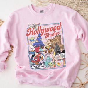 A pink sweatshirt with a colorful graphic design featuring characters from Disney and Hollywood Studios, including Mickey Mouse, Minnie Mouse, and other iconic figures.