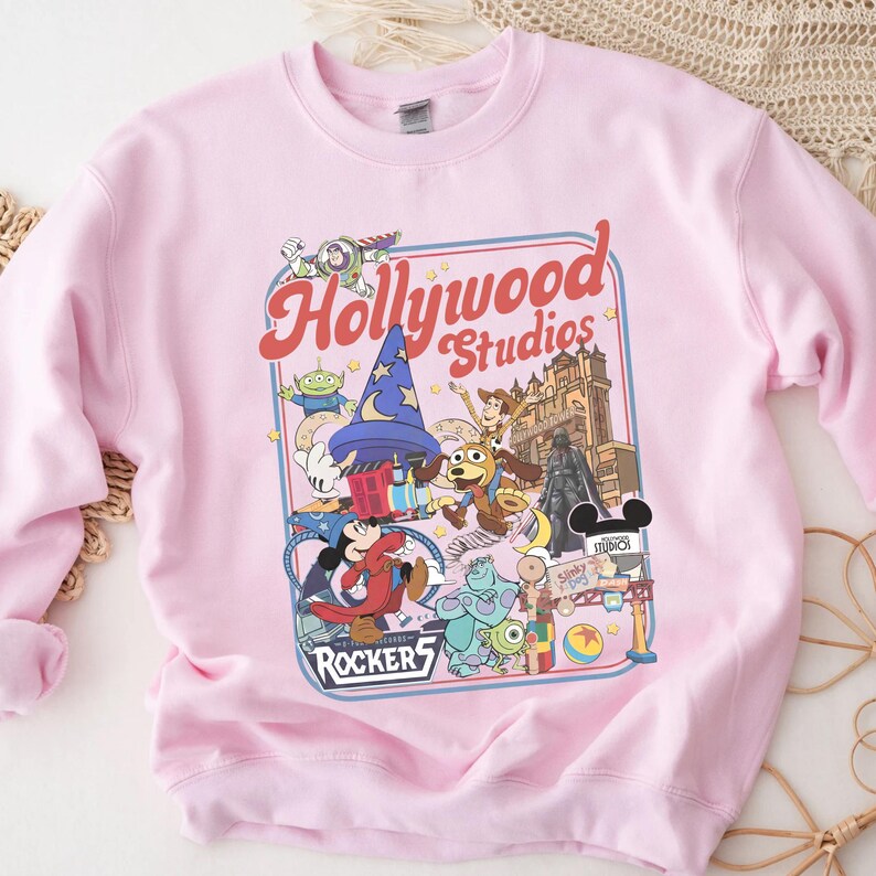 A pink sweatshirt with a colorful graphic design featuring characters from Disney and Hollywood Studios, including Mickey Mouse, Minnie Mouse, and other iconic figures.